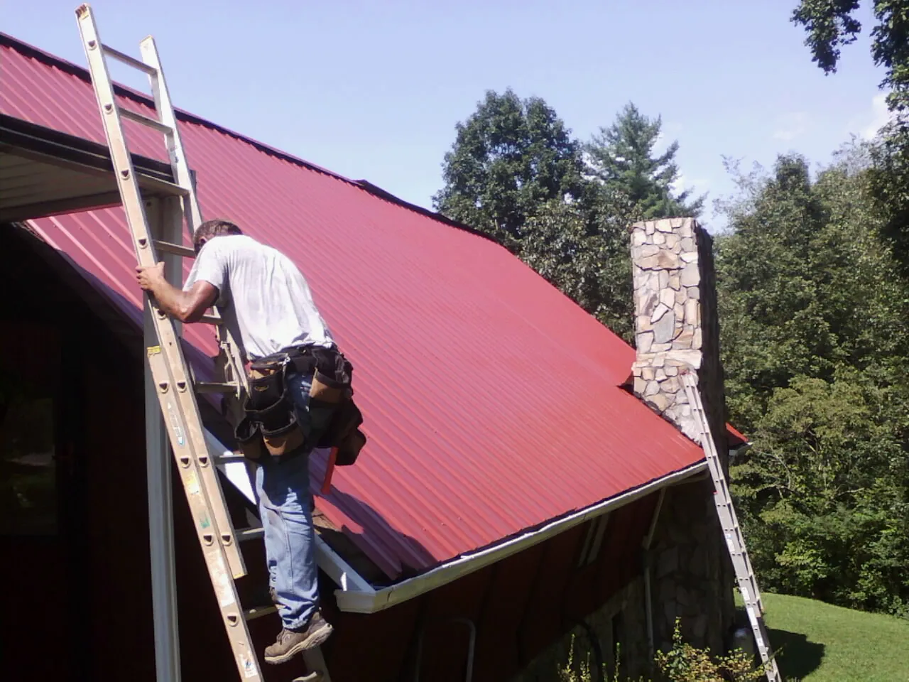 Roofer on a ladder beside a red metal roof and stone chimney — Soffit & Fascia Repair by Roofing Brothers in Pennsylvania Furnace
